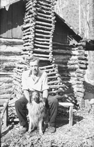 Gene J_ sitting beside chimney with Laddie Boy at little cabin
